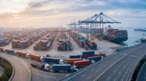 Aerial view of a container shipping terminal with cargo ship, cranes, stacked containers and freight trucks departing